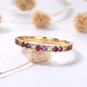 May include: A gold band ring with a row of alternating gemstones in shades of purple, pink, and green. The ring is set on a white surface with blurred floral accents in the background. The ring is a delicate piece of jewelry.