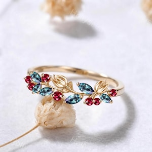 May include: A gold ring featuring a delicate vine design with marquise-cut blue-green stones and round red stones. The band is accented with small, sculpted gold flower buds interspersed among the colorful gemstones in a nature-inspired arrangement.
