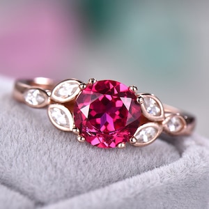 May include: A rose gold ring with a large, round, deep red gemstone surrounded by smaller, clear gemstones. The ring has a delicate, floral design.