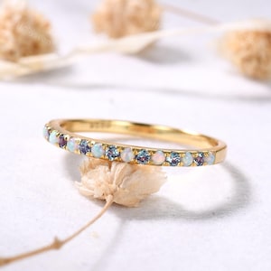 May include: A gold band ring with a row of small, colorful gemstones. The gemstones alternate between white, blue, and purple. The ring is set against a soft, neutral background.