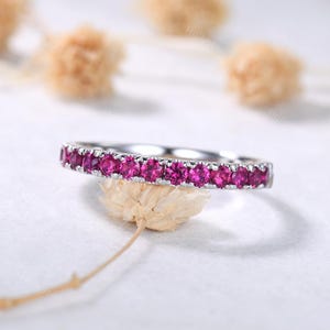May include: A silver ring with a band of vibrant pink gemstones. The ring is set against a soft, neutral background, highlighting the sparkle of the gemstones. The ring is a delicate piece of jewelry.