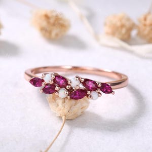 May include: A rose gold ring featuring a cluster of marquise-cut, deep red gemstones and small, round, white gemstones. The ring's band is thin and delicate, with the gemstones arranged in a leaf-like pattern.