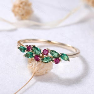 Puede incluir: Un anillo de oro con un grupo de piedras preciosas verdes y rojas. La banda del anillo es de oro pulido, mientras que las piedras preciosas están dispuestas en forma de hoja. Las piedras preciosas son de color verde esmeralda y rojo rubí.