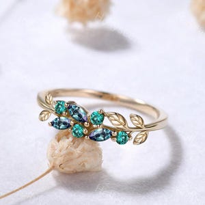 Op de afbeelding: Een goudkleurige ring met een bladmotief, bezet met teal en blauwe edelstenen. De ring heeft een delicate band en is versierd met marquise- en rond geslepen stenen, wat een natuurlijke uitstraling geeft. De edelstenen zijn in een clusterpatroon gerangschikt.