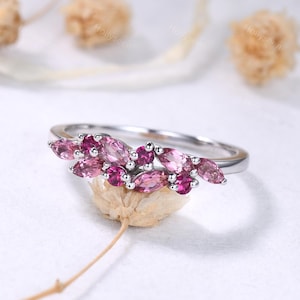May include: A silver ring featuring a cluster of pink gemstones. The ring's design includes marquise-cut and round-cut stones, creating a floral or leaf-like arrangement. The gemstones vary in shades of pink.