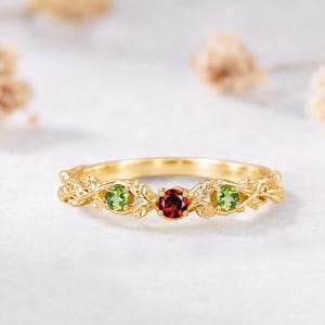 May include: A gold ring featuring a central round red garnet flanked by two round green peridot stones. The band is crafted with a detailed vine and leaf motif that extends across the front, holding the three gemstones in prong settings.