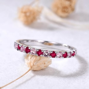 May include: A thin silver band ring featuring a continuous row of alternating round-cut pink and deep red gemstones set in a shared prong setting. The metal band has a polished finish, and the stones are arranged in a repeating pattern around the ring.