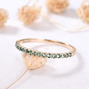 May include: A delicate gold band ring with a row of small, green gemstones. The ring is thin and simple, with a subtle sparkle.