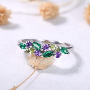 May include: A silver ring featuring a cluster of marquise-cut gemstones in shades of green, purple, and yellow. The gemstones are arranged in a leaf-like pattern, creating a nature-inspired design. The ring is set on a white surface.