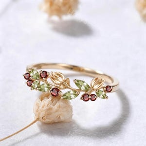 Könnte beinhalten: Ein goldener Ring mit botanischem Design, besetzt mit hellgrünen Steinen im Marquise-Schliff und runden dunkelroten Steinen, die wie Blätter und Beeren angeordnet sind. Die Ringschiene ist schmal und poliert und zeigt ein zartes Zweigmotiv mit zwei kleinen Blütenknospen.