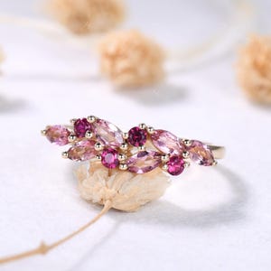 May include: A gold ring featuring a floral design with pink and magenta gemstones. The ring has a delicate arrangement of marquise-cut and round gemstones, creating a cluster of color. The gemstones are set in a gold band.