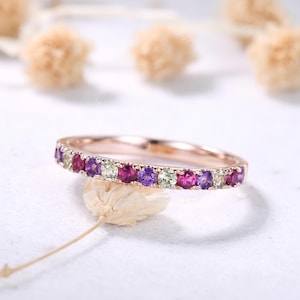 May include: Rose gold band ring with a row of alternating gemstones in shades of purple, pink, and green. The ring is set on a white surface with blurred, light-colored floral accents in the background. A delicate, dried flower stem is in the foreground.