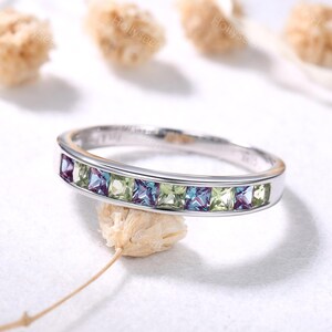 May include: A silver-toned band ring featuring a channel setting with alternating square-cut light green and purple-blue gemstones. The metal band has a polished finish and a smooth, rounded profile, showcasing a series of stones set side by side.