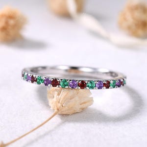 May include: A silver band ring with a row of alternating red, green, and purple gemstones. The ring is set on a white surface with blurred, neutral-toned objects in the background. The ring is a delicate piece of jewelry.