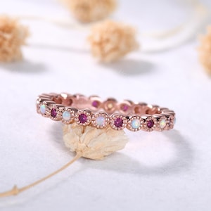 May include: Rose gold ring featuring alternating round-cut gemstones. The ring showcases a pattern of vibrant red rubies and iridescent opals, set within a delicate, textured band. The ring is displayed on a neutral background, highlighting its intricate design.
