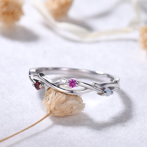 May include: A delicate silver ring with a twisted branch design. The ring features three gemstones: a pink stone, a red stone, and a white opal.