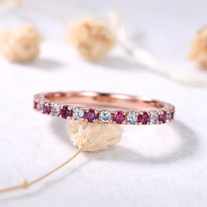 Może przedstawiać: Pierścionek z różowego złota z naprzemiennymi czerwonymi i jasnoniebieskimi kamieniami szlachetnymi. Pierścionek jest osadzony małymi, okrągłymi kamieniami, tworząc ciągły pas koloru. Pierścionek jest prezentowany na białej powierzchni.