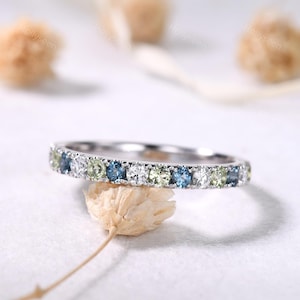 May include: A silver band ring with a row of alternating blue, white, and light green gemstones. The ring is set on a white surface with dried flowers in the background. The ring is a piece of jewelry.