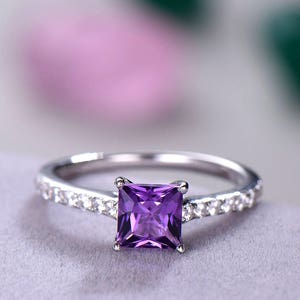 May include: A silver ring featuring a square purple amethyst gemstone, set in a band of small clear gemstones.