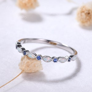 May include: A silver-toned ring featuring an alternating pattern of marquise-cut iridescent opals and small round blue gemstones set along the band. The metal band has a polished finish and a slim profile, showcasing a delicate and symmetrical design.