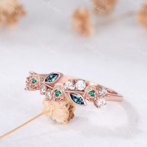 Op de afbeelding: Roségouden ring met een bloemmotief, teal en witte edelstenen. De ring heeft kleine roosvormige accenten, teal en blauwe bladvormige stenen en witte ronde stenen. De ring ligt op een witte ondergrond.