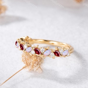 May include: A gold band ring featuring an alternating pattern of marquise-cut red rubies, oval-cut opals, and round-cut clear white diamonds. The gemstones are set in a continuous row along the top half of the polished yellow gold metal band.