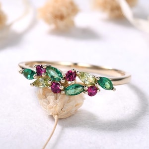 May include: A gold ring featuring a cluster of green, light green, and magenta gemstones. The gemstones are arranged in a leaf-like pattern across the top of the ring. The ring is set against a soft, white background.