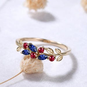 May include: A gold-colored ring with a leaf design. The ring features red and blue gemstones. The gemstones are arranged in a pattern that resembles leaves and berries. The ring is set on a white surface.