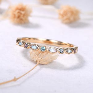 May include: A gold band ring with alternating oval and round gemstones in shades of blue and purple. The ring is set on a white surface with a blurred background.
