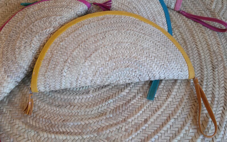 May include: A half-circle straw clutch with a yellow trim and a brown leather strap. The clutch is made of woven straw and has a teal blue leather strap.