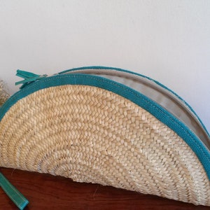 May include: A half-circle woven straw clutch bag with a teal zipper and trim. The bag is open and the lining is visible.