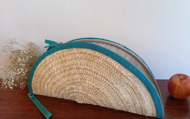 May include: A half-circle woven straw clutch bag with a teal zipper and trim. The bag is open and the lining is visible.