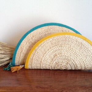 May include: Two half-circle straw clutches with a woven design. The larger clutch has a teal trim and the smaller clutch has a yellow trim. Both clutches have a brown leather strap.