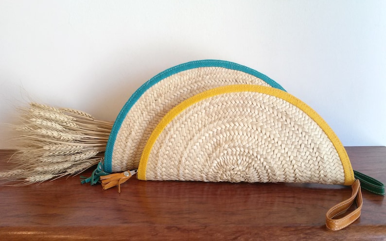 May include: Two half-circle straw clutches with a woven design. The larger clutch has a teal trim and the smaller clutch has a yellow trim. Both clutches have a brown leather strap.