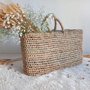 May include: A rectangular woven straw tote bag with two handles, filled with wheat stalks and white baby's breath flowers. The bag is a natural beige colour and sits on a white textured surface.