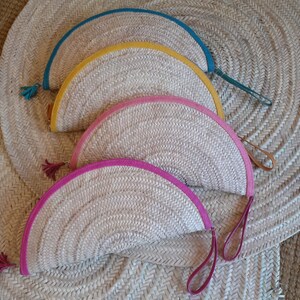 May include: Four half-circle woven straw purses with leather handles and colored leather trim. The purses are arranged in a stack, with the top purse having a teal trim, the next having a yellow trim, the next having a pink trim, and the bottom purse having a pink trim.
