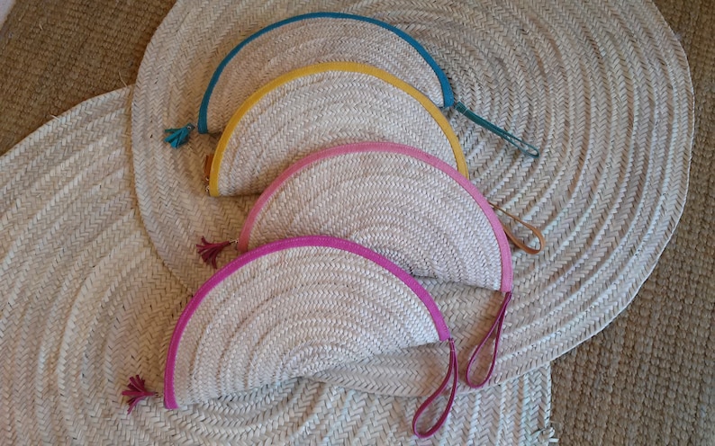 May include: Four half-circle woven straw purses with leather handles and colored leather trim. The purses are arranged in a stack, with the top purse having a teal trim, the next having a yellow trim, the next having a pink trim, and the bottom purse having a pink trim.