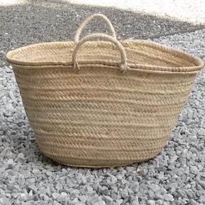 May include: A large, natural woven basket with two handles. The basket is made of straw and has a light brown color. The basket is sitting on a bed of small, gray stones.