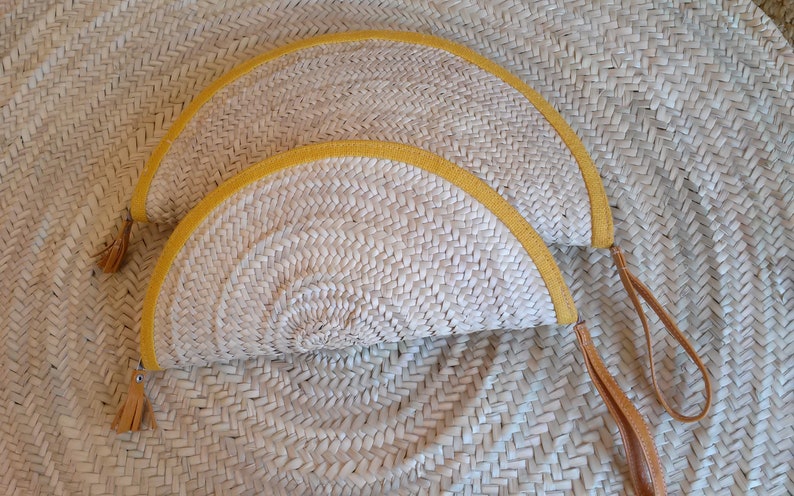 May include: Two half-circle straw purses with yellow trim and brown leather straps. The purses are woven and have a natural straw color.