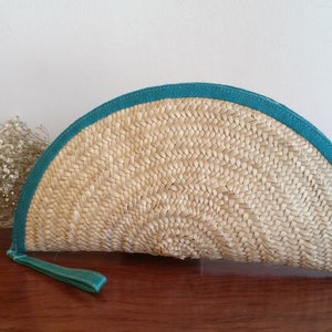 May include: A half-circle shaped straw clutch with a teal blue trim. The clutch has a woven texture and a tassel.