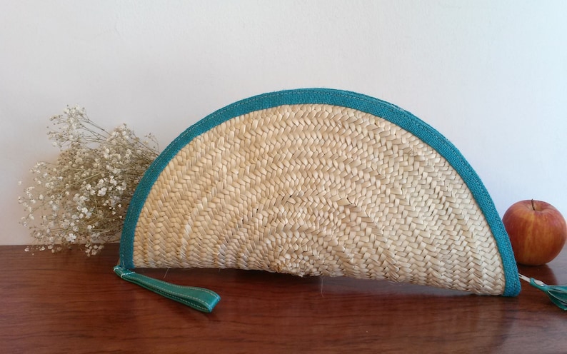 May include: A half-circle shaped straw clutch with a teal blue trim. The clutch has a woven texture and a tassel.