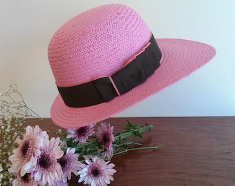 Könnte beinhalten: Ein rosa gewebter Sonnenhut mit breiter Krempe und einem dunkelbraunen Band. Der Hut wird mit einem Strauß rosa und lila Blumen präsentiert, was eine sommerliche und feminine Ästhetik erzeugt.