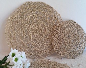 May include: Two round woven straw placemats, one larger than the other, with a natural beige color. The placemats are made of a woven material that creates a delicate, intricate pattern.