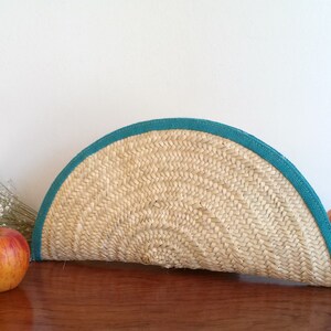 May include: A half-circle straw clutch bag with a teal blue trim and a tassel. The bag is sitting on a wooden surface with apples nearby.