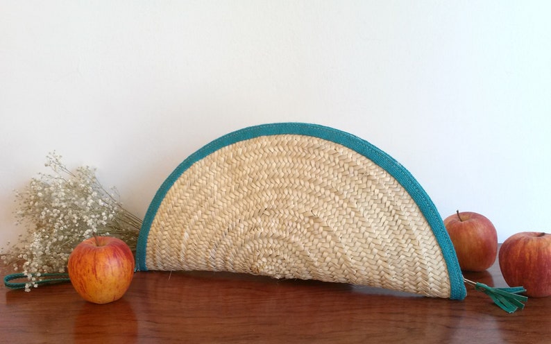 May include: A half-circle straw clutch bag with a teal blue trim and a tassel. The bag is sitting on a wooden surface with apples nearby.