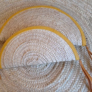 May include: Two half-circle straw purses with yellow trim and brown leather straps. The purses are woven and have a natural straw color.