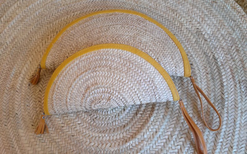 May include: Two half-circle straw purses with yellow trim and brown leather straps. The purses are woven and have a natural straw color.