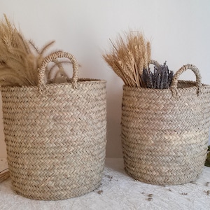 Może przedstawiać: Dwa plecione kosze z uchwytami, wypełnione suszoną pszenicą i lawendą. Kosze są cylindryczne i wykonane z jasnobrązowego, tkanego materiału. Tło to biała ściana i biała powierzchnia.