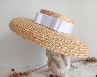 May include: A wide-brimmed straw hat with a flat crown and a wide, circular brim. The hat features a smooth, white ribbon band wrapped around the base of the crown, tied into a structured bow at the front, creating a clean and simple aesthetic.