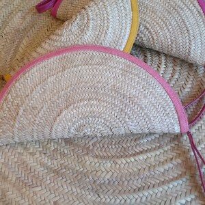 May include: Two half-circle woven straw bags with pink and yellow trim and leather handles. The bags are lying on a woven straw surface.
